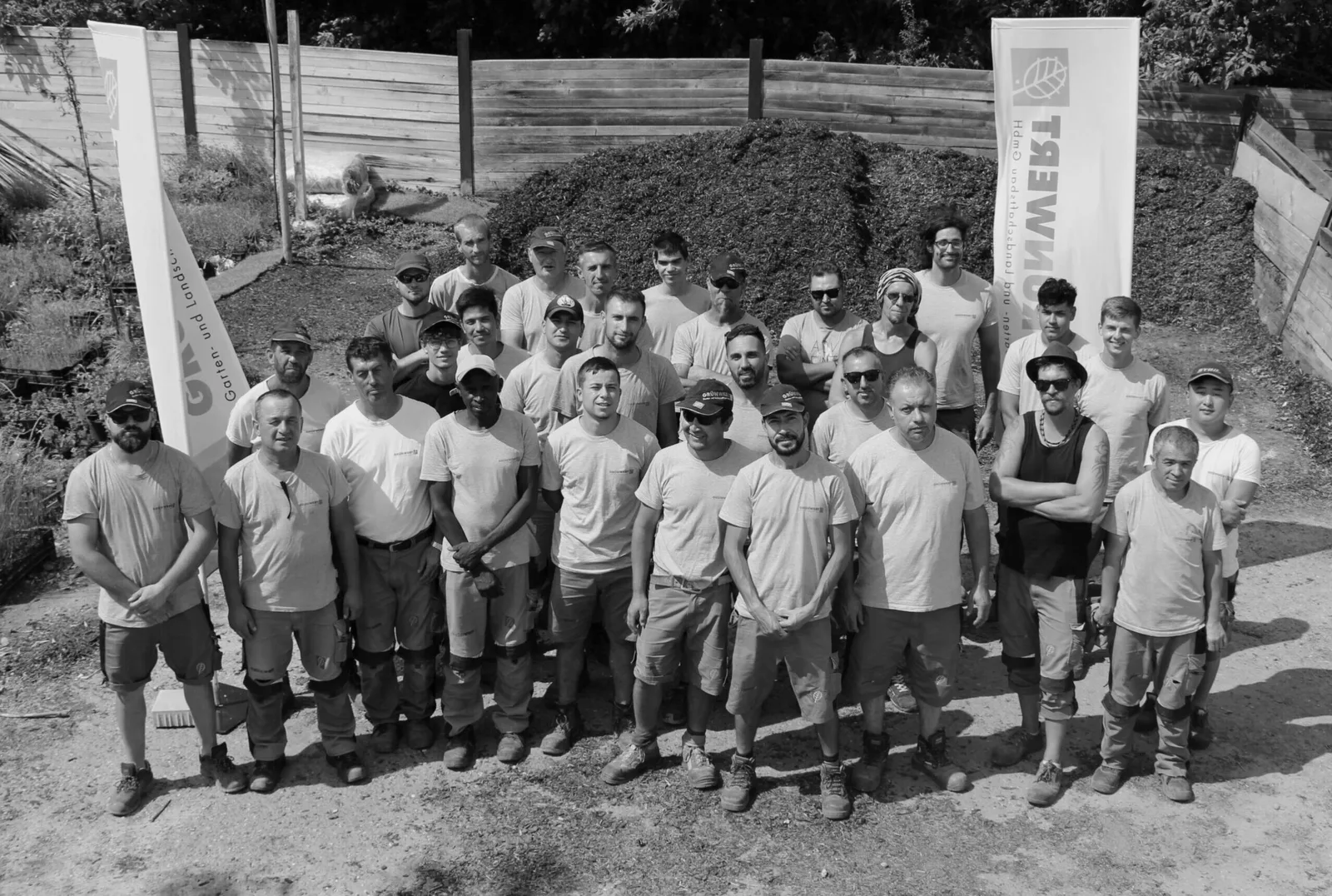 Gruppenfoto eines Grünwert-Teams vor einem Erdhaufen, mit Grünwert-Fahnen, schwarzweiß